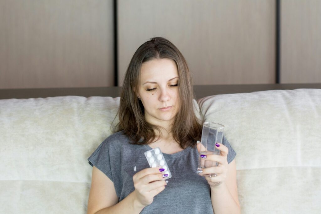 A white woman looks tired and pale she is sitting on a white sofa holding medications.